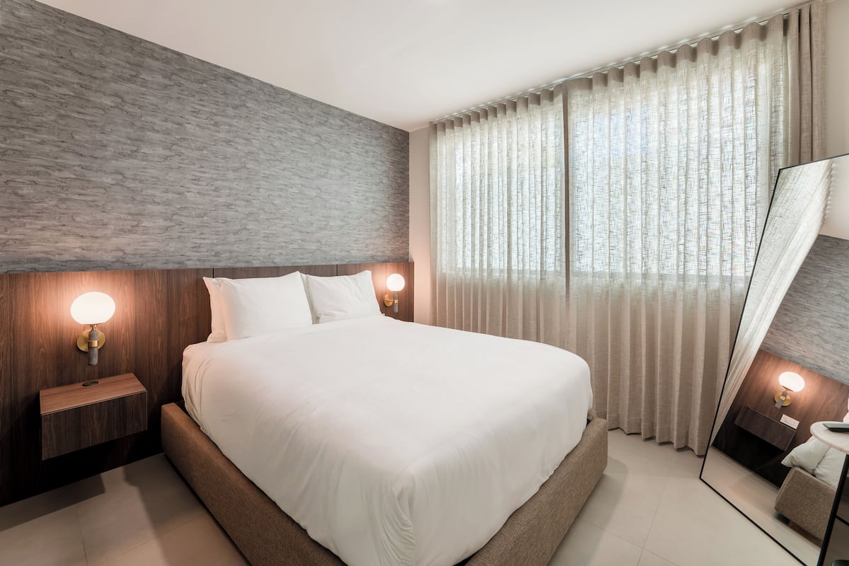 A modern hotel room with a large bed, wooden headboard, two bedside lamps, a large mirror, and a window with sheer curtains.