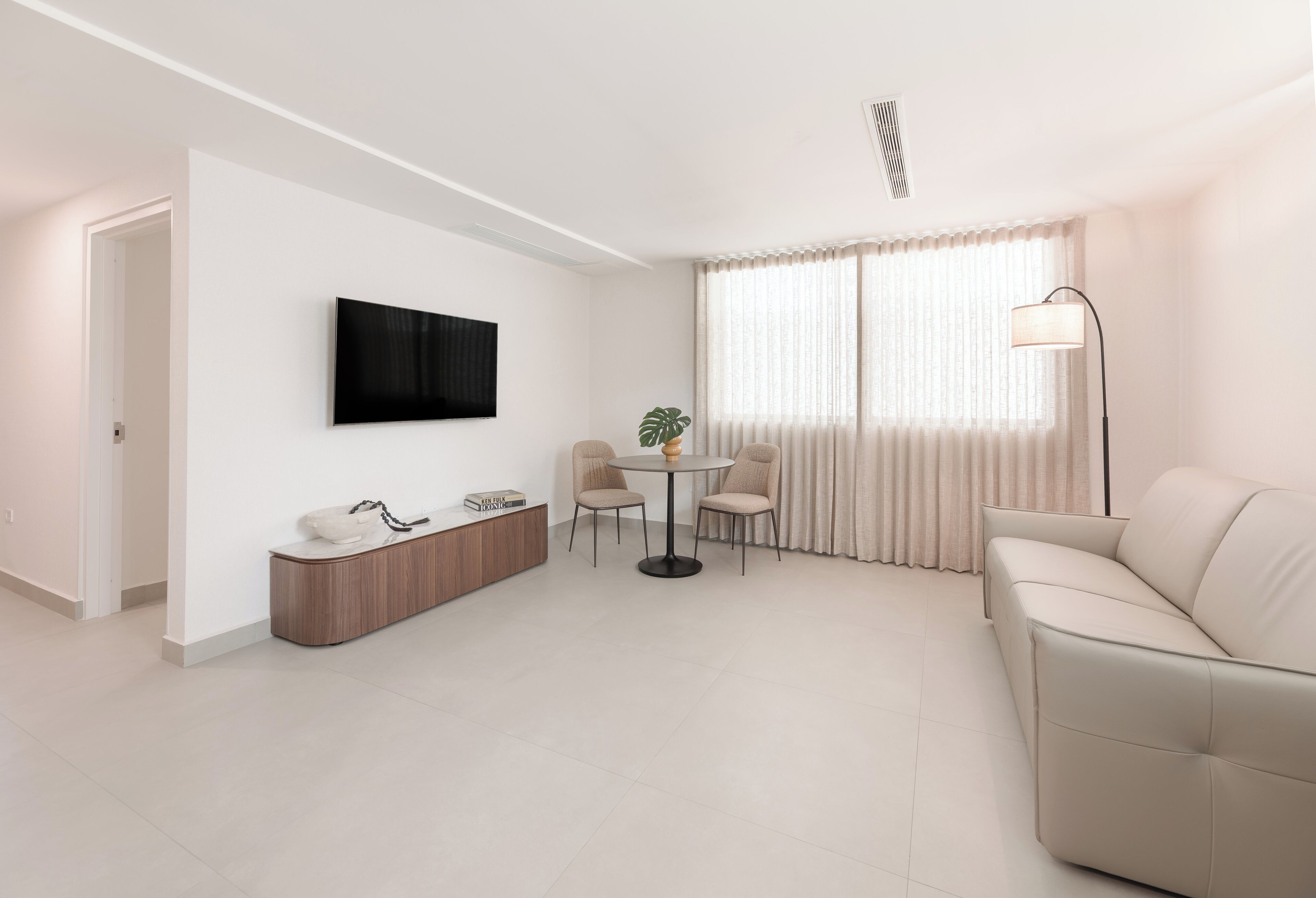 A modern living room with a sofa, a TV mounted on the wall, a dining table with chairs, and a lamp.