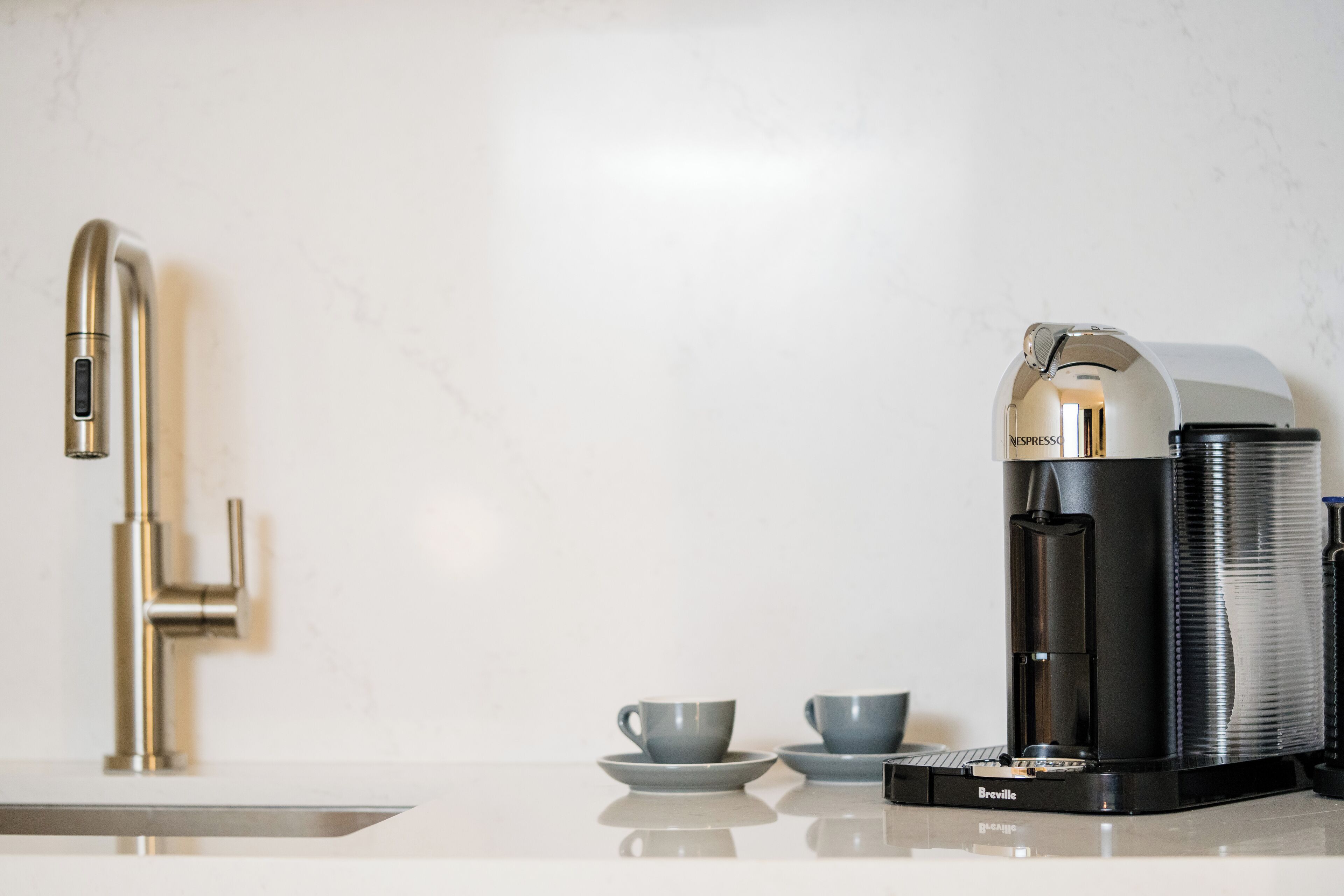 A Breville coffee maker on a white countertop with a modern faucet and two cups.