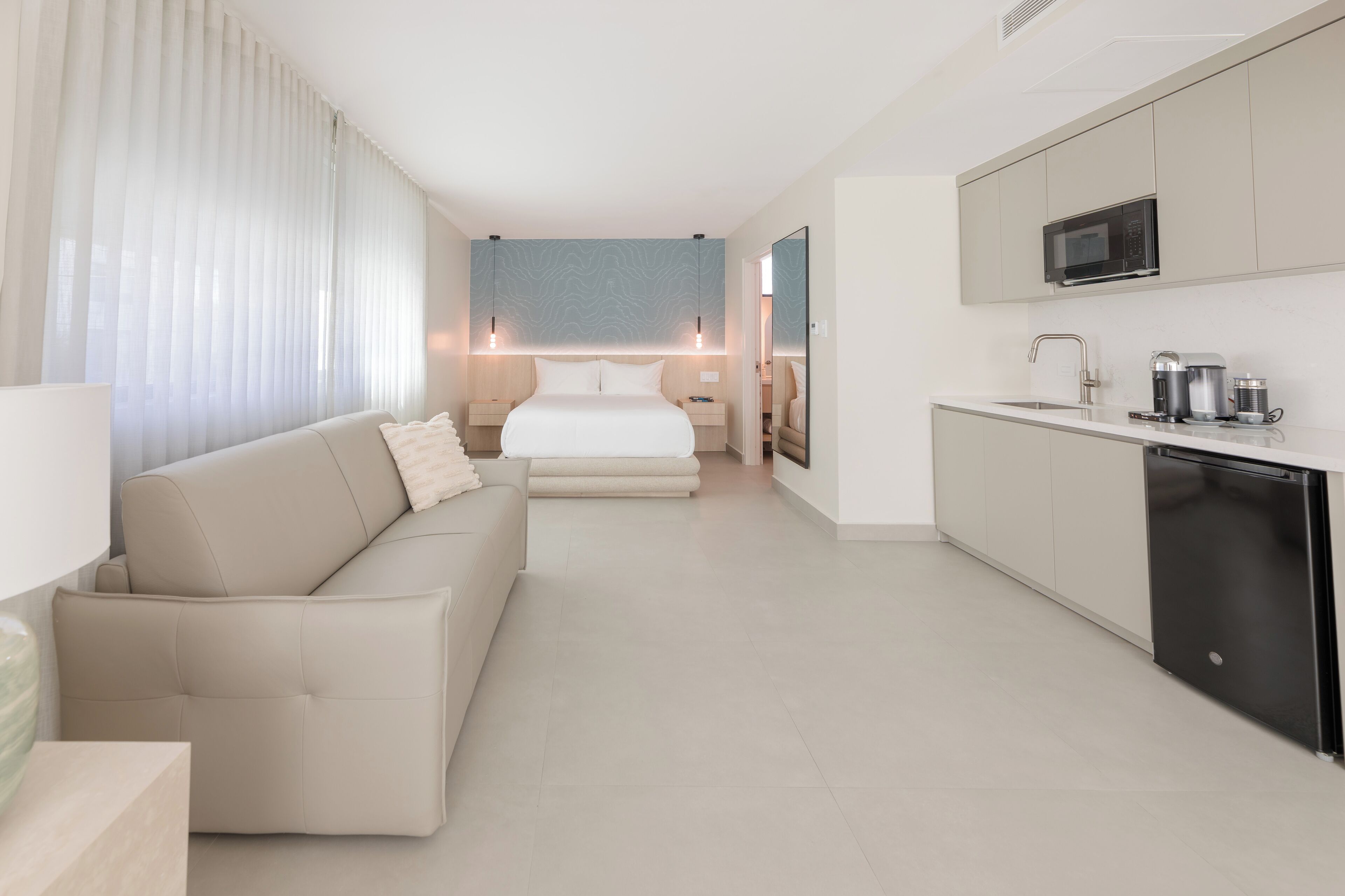 A modern hotel room with a sofa, a bed, a kitchenette, and a large window with sheer curtains.