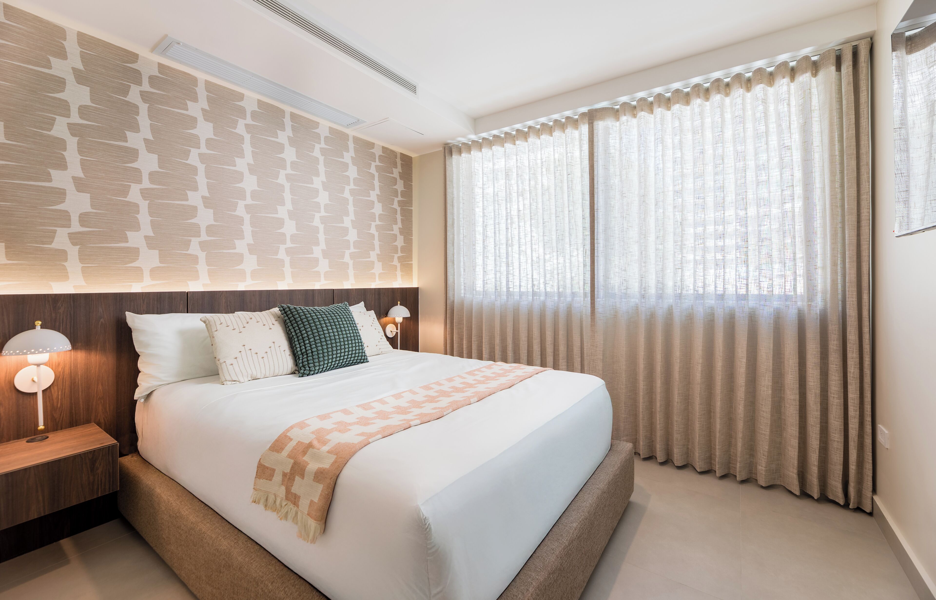 A modern hotel room with a large bed, a headboard with a textured pattern, a bedside lamp, and a window with sheer curtains.
