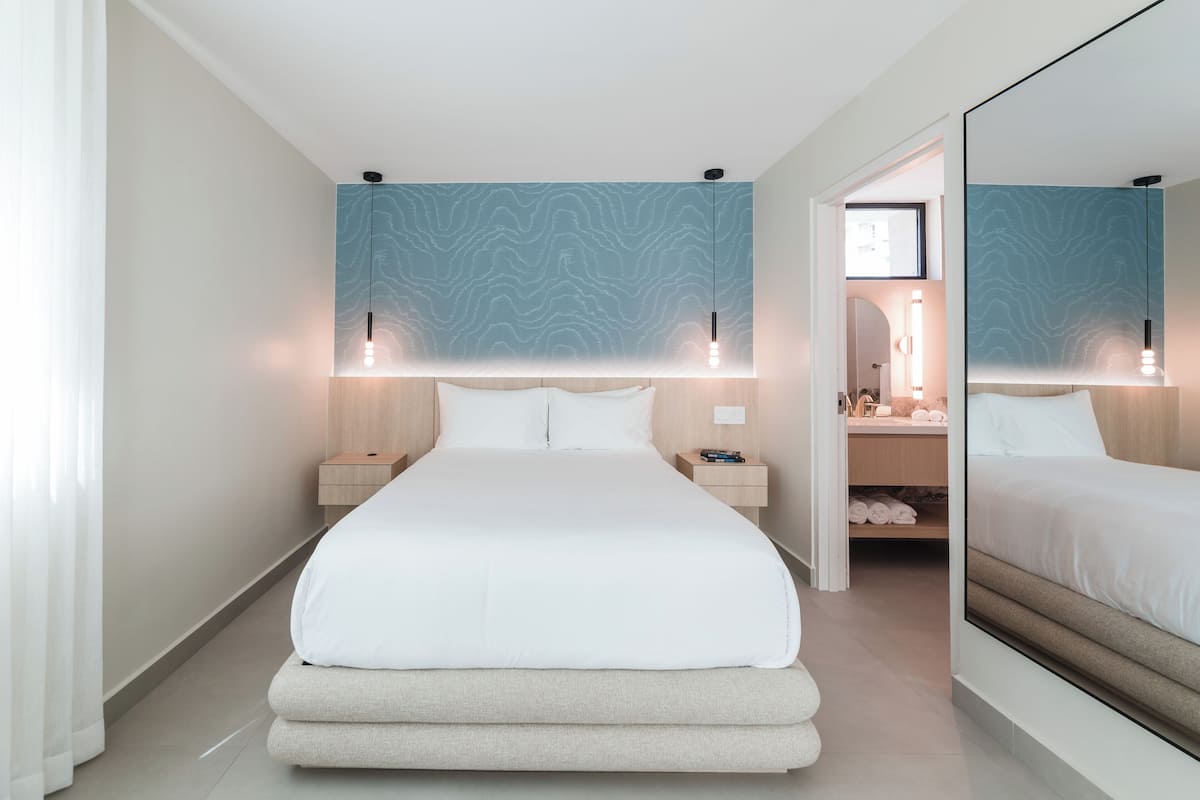 A modern hotel room with a large bed, bedside tables, a mirror, and a bathroom visible through an open door.