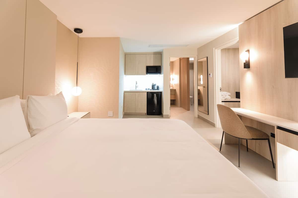 A hotel room with a large bed, a desk, a chair, a kitchenette, and a bathroom visible through an open door.
