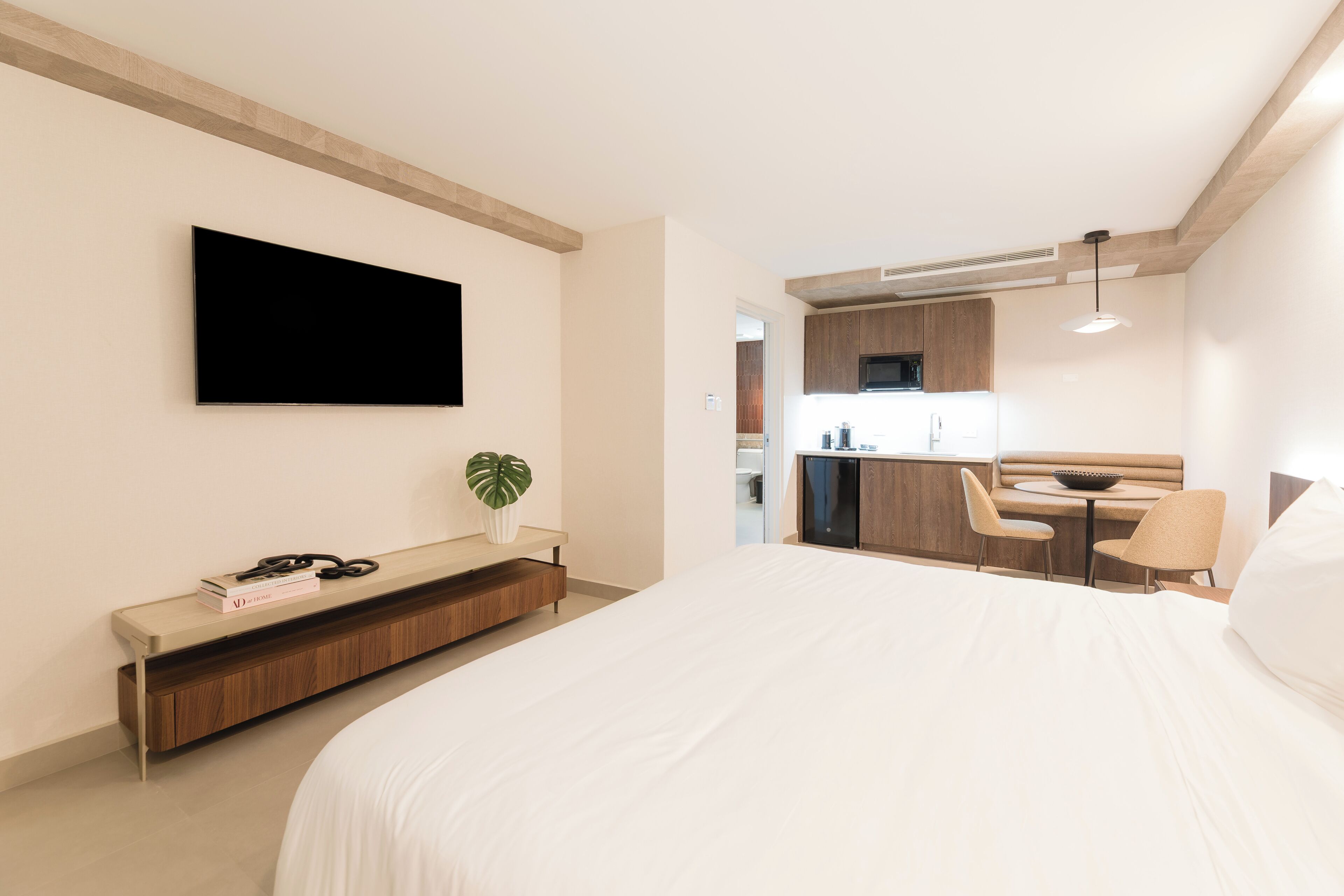 Una habitación de hotel moderna con una cama grande, un televisor de pantalla plana montado en la pared, una pequeña zona de estar con una mesa y sillas, y una kitchenette al fondo.