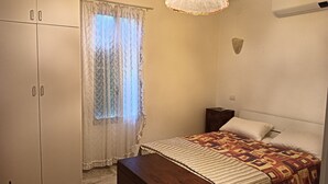 Un dormitorio con cama, mesita de noche, cómoda, ventana con cortinas translúcidas y lámpara en el techo.