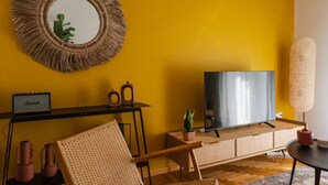 Un soggiorno con una parete gialla, uno specchio rotondo, una televisione a schermo piatto, un tavolino consolle in legno, una sedia intrecciata e una piccola pianta in vaso.