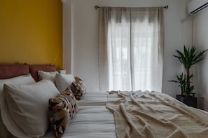 A bedroom with a bed, pillows, a blanket, a window with curtains, and a potted plant. - Urban Comfort - 2BR Apartment in Koukaki (Athina)