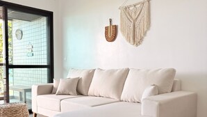 A modern living room with a beige sectional sofa, a woven ottoman, a macrame wall hanging, and a view through a large window. - Ocean View of Recreio Beach Lc103 (Rio de Janeiro)