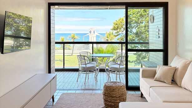 A modern living room with a large sliding glass door leading to a balcony with a view of the sea and a distant tower. - Ocean View of Recreio Beach Lc103 (Rio de Janeiro)