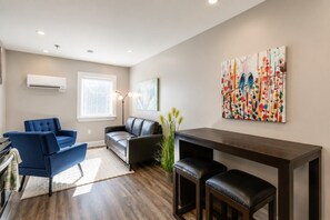 Una sala de estar moderna con un sillón azul, un sofá de cuero negro, una mesa de centro de madera y un cuadro abstracto colorido en la pared.