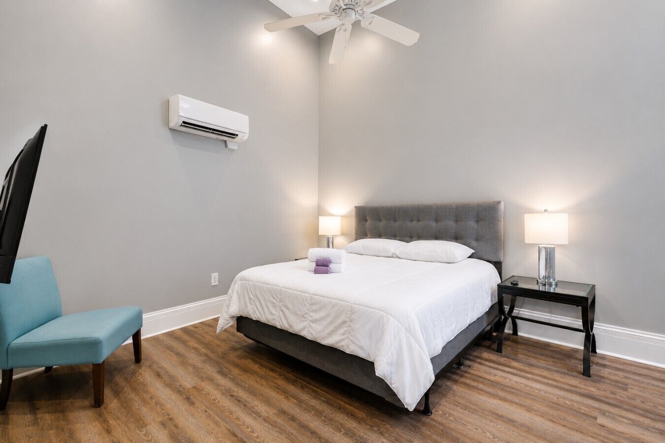 A bedroom with a bed, a chair, a nightstand, a lamp, an air conditioner, and a ceiling fan.