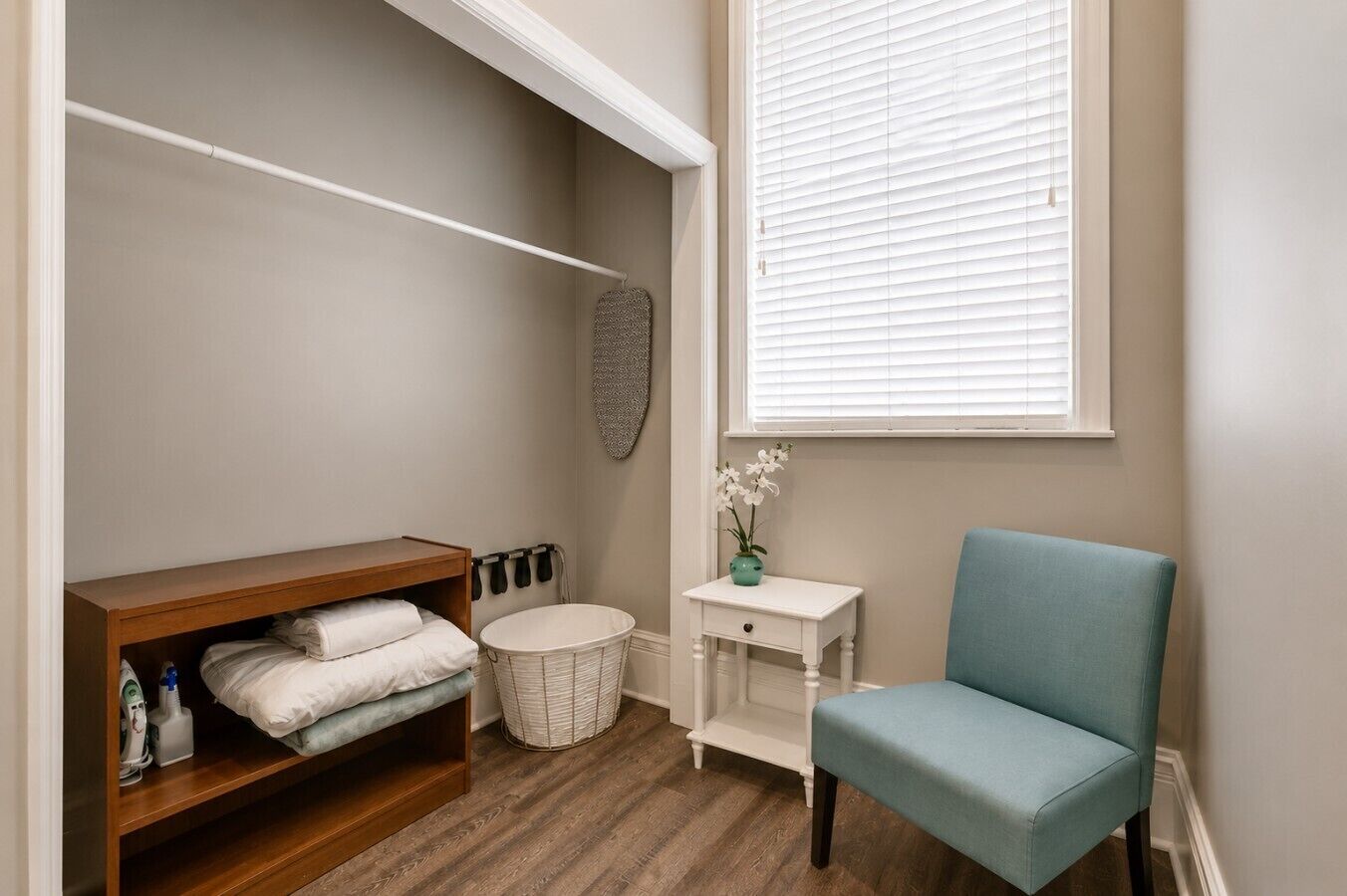 Une salle de bain avec une étagère en bois, une chaise blanche, une petite table blanche et une fenêtre avec des stores.