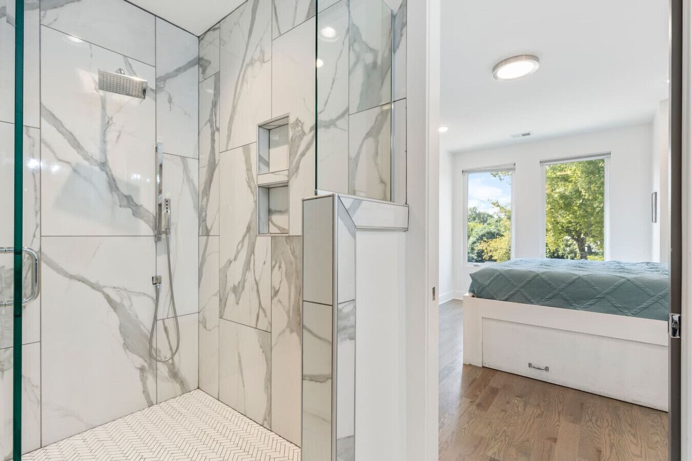 A modern bathroom with marble tiles, a shower, and a bedroom with a window and a bed.