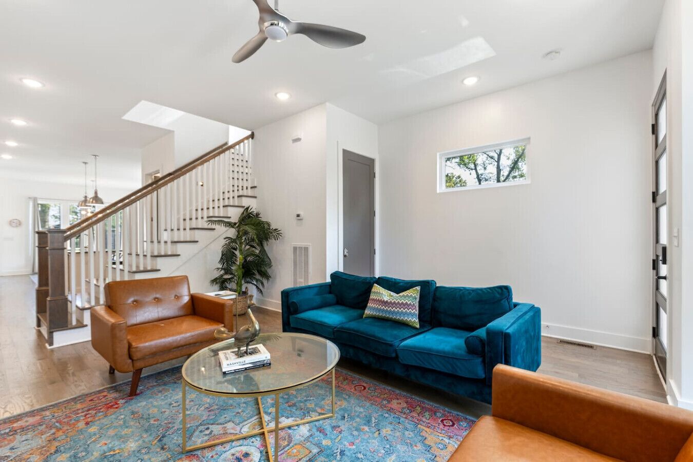 A spacious living room with a blue sofa, a brown leather armchair, and a glass coffee table. A staircase with a wooden railing leads to another level.