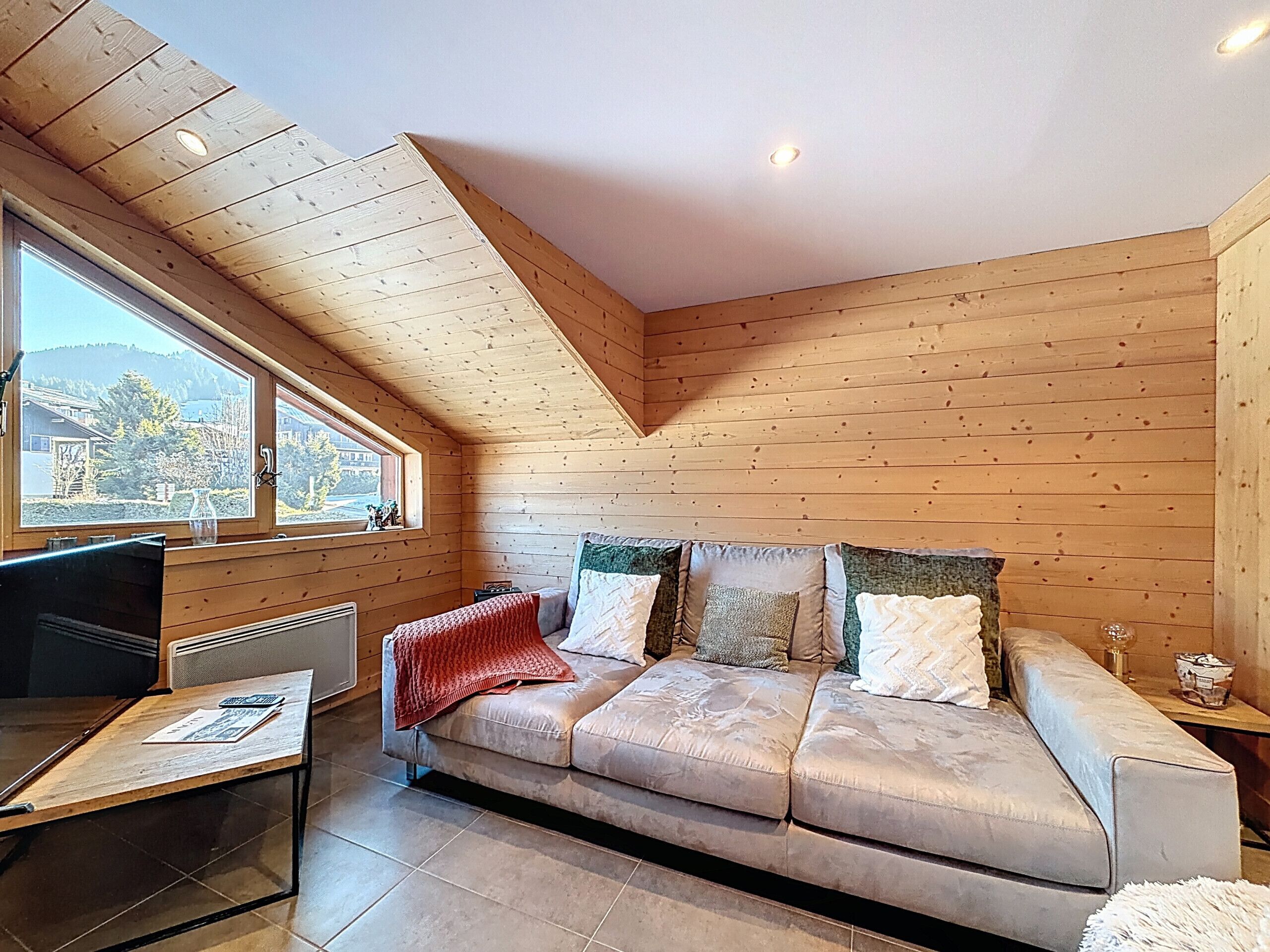 Ein gemütliches Zimmer mit Holzbalkendecke, einem großen Fenster, einer Couch mit vielen Kissen, einem kleinen Tisch und einem Heizkörper.