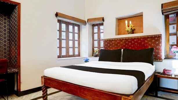 Una camera da letto con un letto in legno, due finestre con stipiti in legno, un comodino con un telefono e una mensola a muro con un vaso.