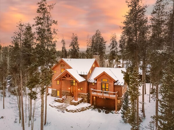 A log cabin with a stone foundation, wooden exterior, and a snow-covered roof, surrounded by tall pine trees.