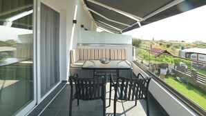 Un balcón con puerta de vidrio, un banco acolchado, una mesa con sillas y vistas al paisaje circundante.