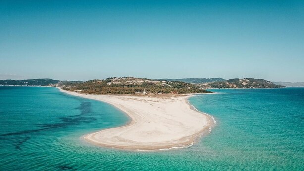 Vue aérienne d’une plage aux eaux turquoise et limpides, bordée d’un rivage sablonneux, avec un paysage vallonné en arrière-plan.