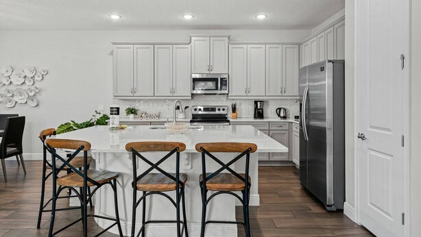 A modern kitchen with white cabinets, a stainless steel refrigerator, a microwave, and a large island with bar stools. - Last Minute Disney Getaway! Luxury Villa with Private Pool, Game Room & Themed Bedrooms. Book Now! #161617 (Orlando)