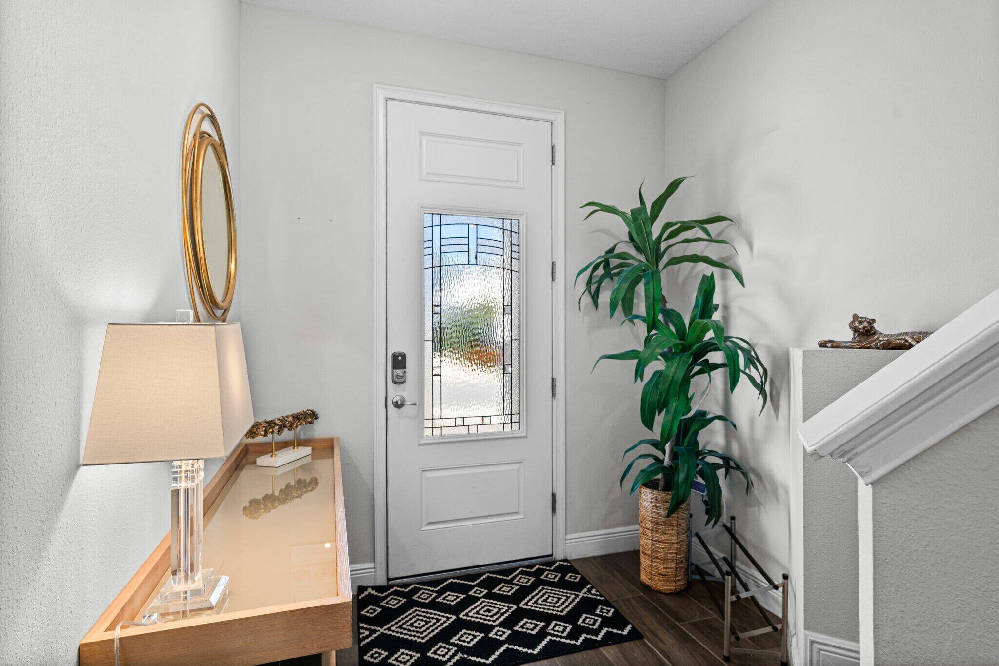 Une porte blanche avec un panneau de verre, une lampe en verre transparent, un tapis à motifs et une grande plante verte.