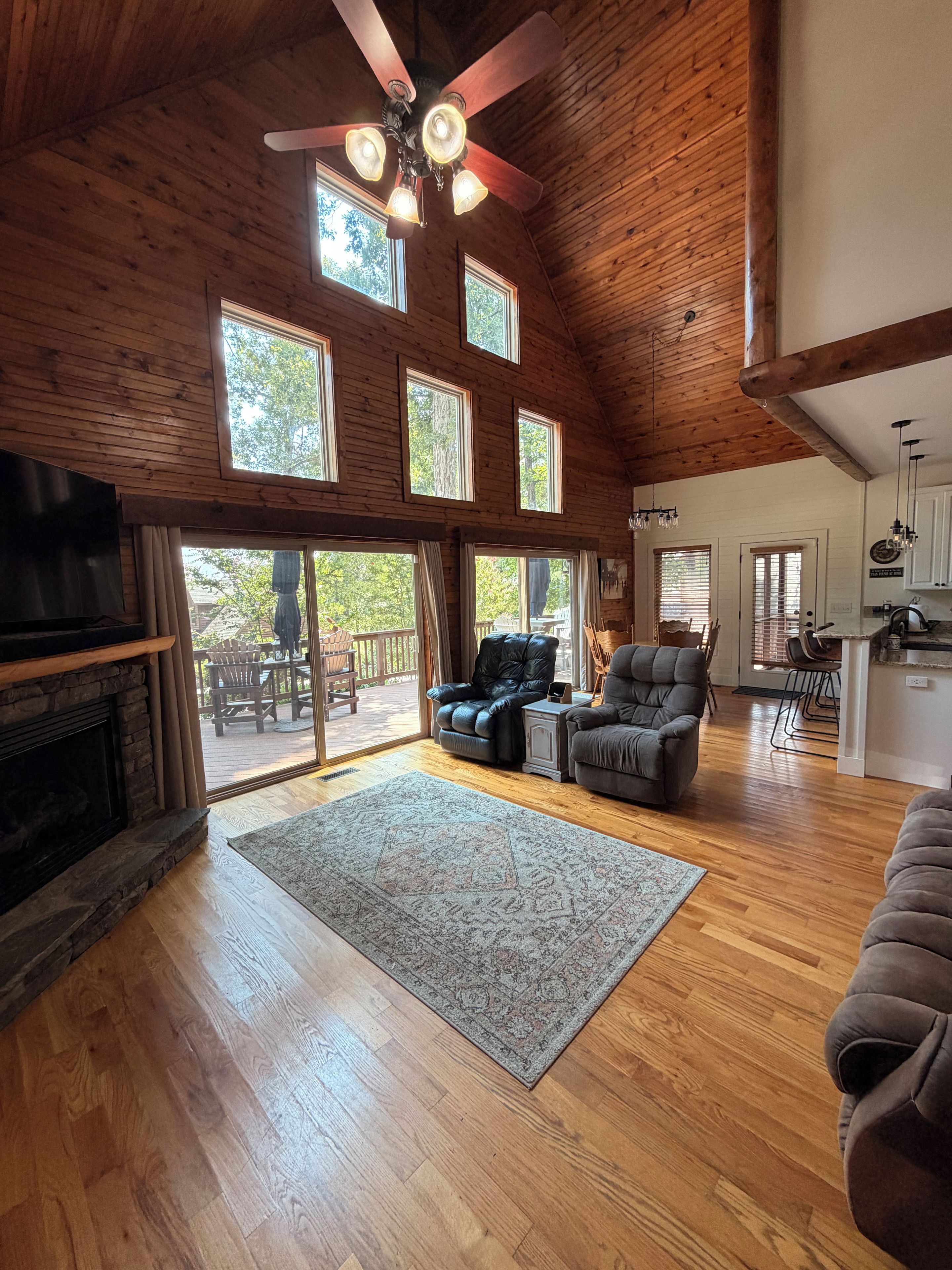 A spacious living area with a fireplace, a ceiling fan, and a large window overlooking a deck.