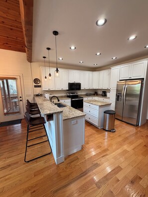 A modern kitchen with white cabinets, a stainless steel refrigerator, a built-in oven, and a granite countertop island. - Peaceful Lake Keowee Home, Quiet Cove in Serenity Bay, Great for Clemson Events (Seneca)