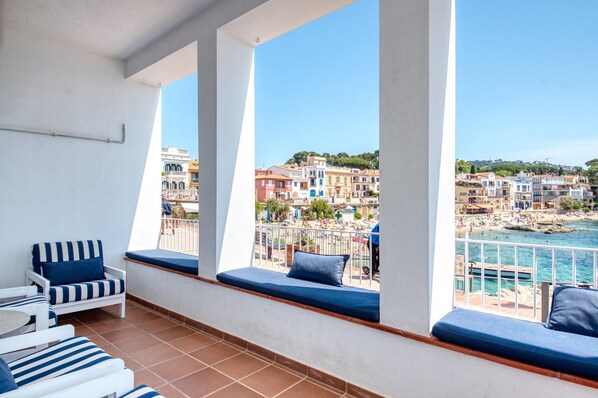 A balcony with white columns, blue cushioned seating, and a view of a coastal town. - House on the Seafront in Calella – Iconic “Casa Rosa” (Calella de Palafrugell)