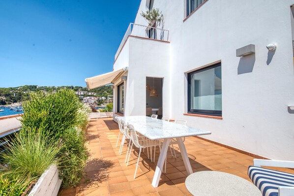 A modern outdoor patio with a white table and chairs, a view of the sea, and a white building with large windows. - House on the Seafront in Calella – Iconic “Casa Rosa” (Calella de Palafrugell)