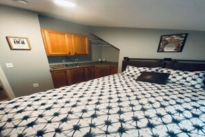 A bedroom with a bed, wooden cabinets, a kitchen area, and a framed picture on the wall.