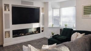Un salon moderne doté d’un grand téléviseur à écran plat fixé au mur, d’une cheminée en dessous, d’un canapé vert et d’une fenêtre avec des stores.