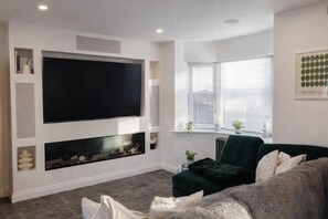 A modern living room with a large flat-screen TV mounted on the wall, a fireplace below it, a green sofa, and a window with blinds. - Hot tub - Bbq - Garden - Parking - Sleeps 8 (Worcester)