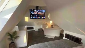 Une chambre moderne aménagée dans les combles, dotée d’un puits de lumière, d’un grand téléviseur à écran plat, d’une baignoire intégrée et d’une petite commode blanche.