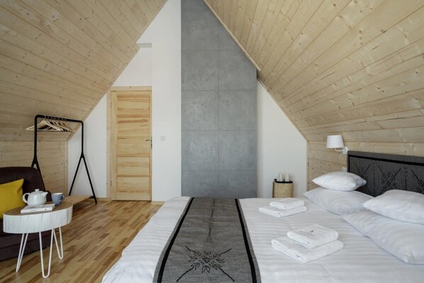 A bedroom with a sloped ceiling, a bed with white linens, a bedside table, a small round table with a kettle, and a wooden coat rack.