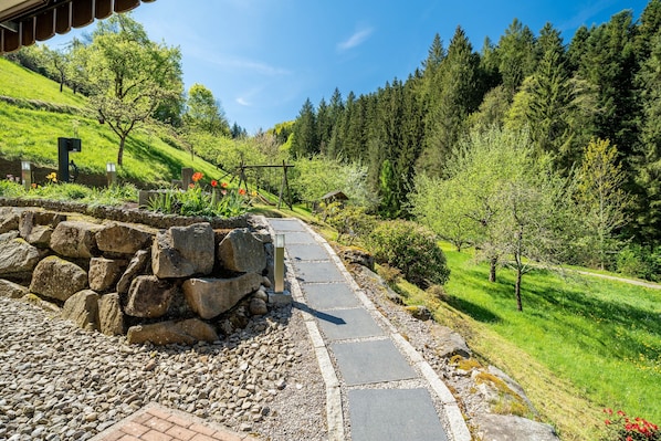 Ein steiniger Weg führt bergauf, im Hintergrund ein Holthaus und eine kleine Werkstatt.