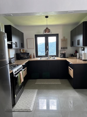 Una cocina moderna con armarios oscuros, un refrigerador de acero inoxidable, un microondas, una estufa y un amplio ventanal con vistas a las montañas.
