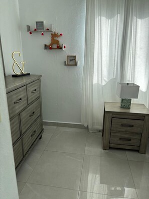 A room with a wooden dresser, a nightstand, and a wall-mounted shelf with decorative items.