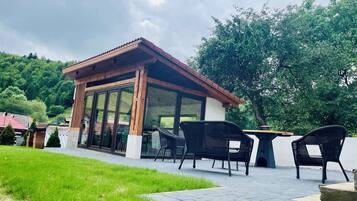 A covered outdoor seating area with a table and chairs, surrounded by a grassy lawn and a paved walkway.