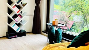 Ein Zimmer mit großem Fenster, einem weißen Bücherregal, einem kleinen runden Tisch mit Kerzen und einem gelben Sofa.