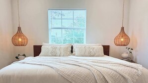A neatly made bed with a white comforter and two decorative pillows, a wicker chair, and a bedside table with a small plant. - poolside getaway w/hot tub & relaxing courtyard (Houston)