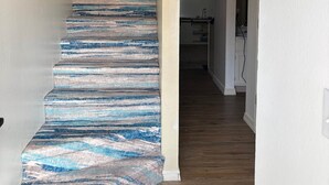 Un escalier recouvert d’un tapis à rayures bleues et blanches, un mur blanc orné d’un panneau indiquant « BE », et un coin cuisine à l’arrière-plan.