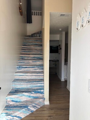 Una escalera con un suelo a rayas azul y blanco, una pared blanca con un cartel que dice 'BE' y una zona de cocina al fondo.