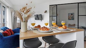 Una sala de estar moderna con un sofá azul, una mesa de comedor con cruasanes y jugo de naranja, y un televisor en la pared.