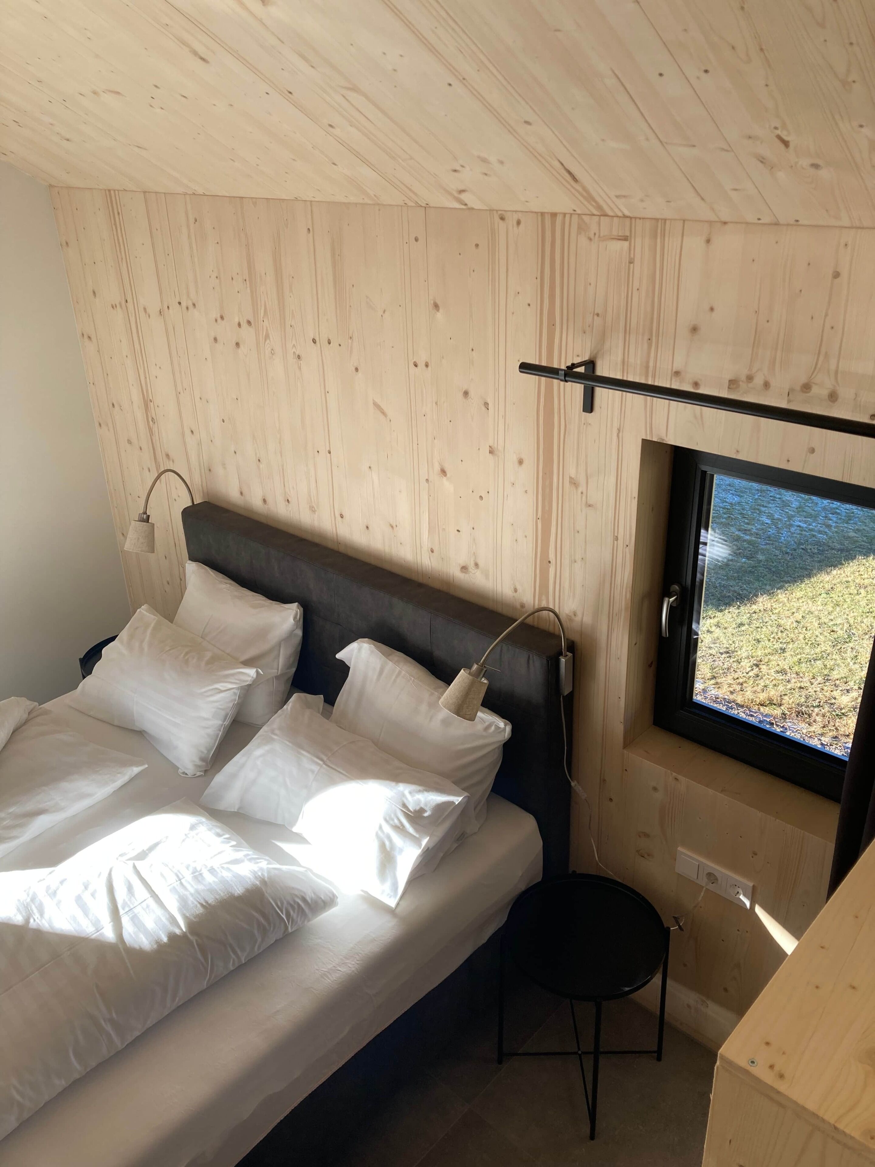 A bedroom with a bed, pillows, a bedside lamp, a window with a view, and a small stool.