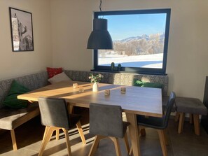 A dining area with a wooden table, grey upholstered chairs, a bench with cushions, and a large window with a view of a snowy landscape. - Alp Chalet Gundersheim (Gundersheim)