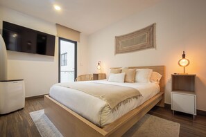 A modern bedroom with a large bed, a flat-screen TV mounted on the wall, a bedside table with a lamp, and a small refrigerator.