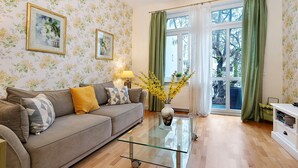 Una sala de estar con un sofá, una mesa de centro de vidrio, un jarrón con flores y una ventana con cortinas.