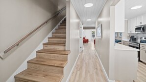 A modern interior with a wooden staircase, a kitchen with stainless steel appliances, and a hallway leading to a dining area. - Beach Bear Townhome: Steps to Sand & Schooners, Pet Friendly, Private Yard (Panama City Beach)
