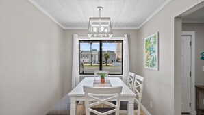A dining area with a table, chairs, a hanging light fixture, a window with a view of buildings, and a door leading to another room. - Beach Bear Townhome: Steps to Sand & Schooners, Pet Friendly, Private Yard (Panama City Beach)
