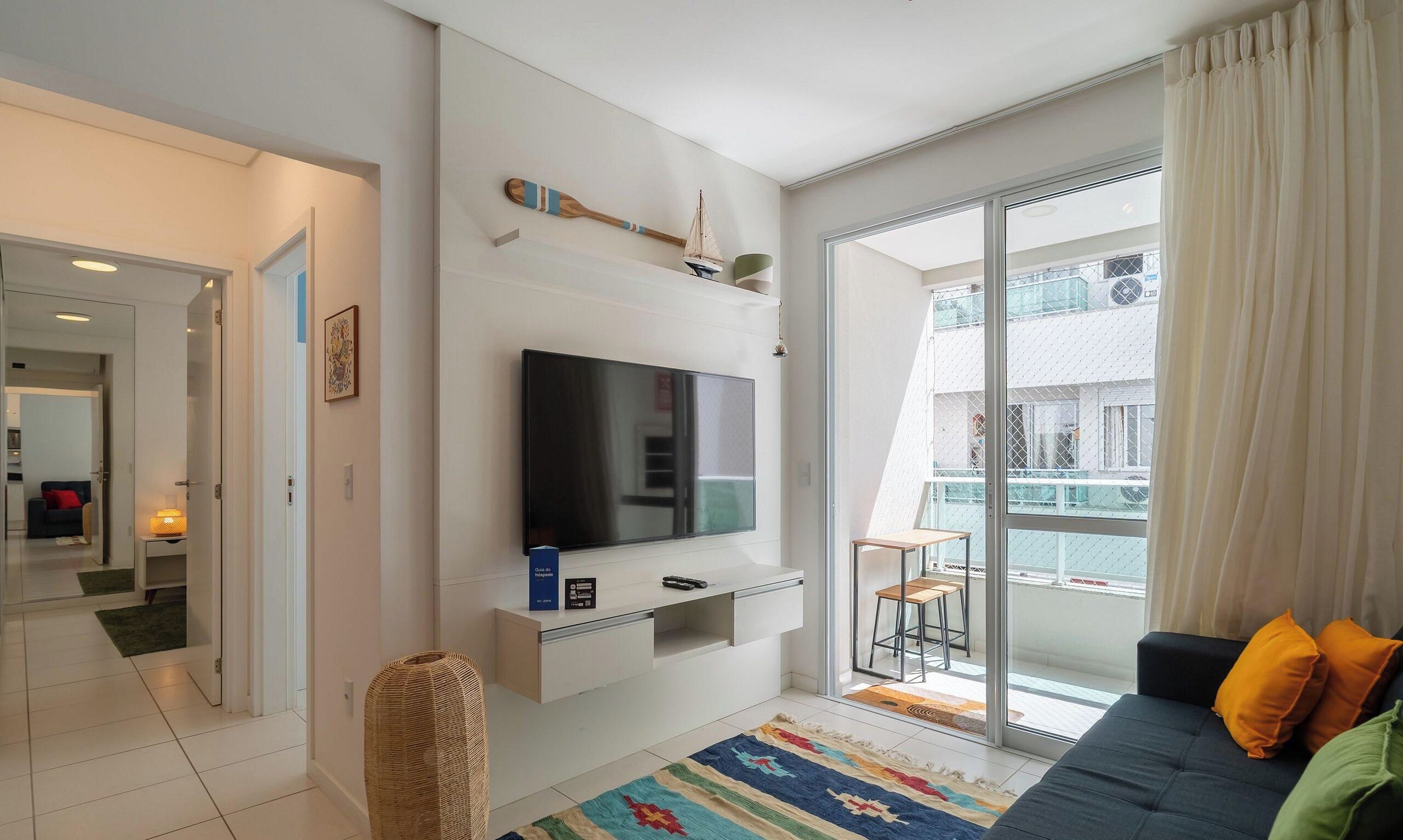 A modern living room with a flat-screen TV, a sofa, a small table, and a balcony with a view.